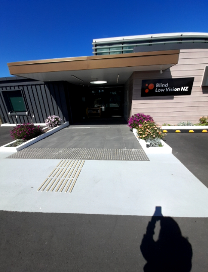 Level concrete path leads to the Blind Low Vision NZ entrance. It features light-coloured directional and warning tactile ground surface indicators, flanked by flower beds. Dark asphalt driveway is...