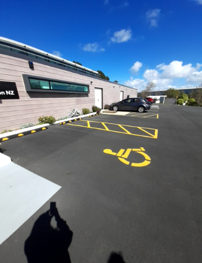 Accessible parking bay with yellow wheelchair symbol on black asphalt. A level concrete pathway leads to the building's white entrance doors. Black and yellow wheel stops border garden beds.
