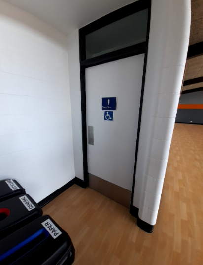 White door with a silver vertical pull handle, an accessible toilet symbol, and male figure sign. Level light timber floor, black recycling bins partially visible on the left.