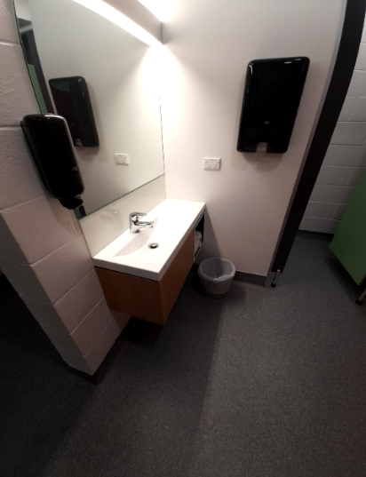 Washroom with level grey textured floor, white walls and bright overhead lighting. Features a sink, tap, counter, wall-mounted soap & hand drying dispensers, plus a small bin.