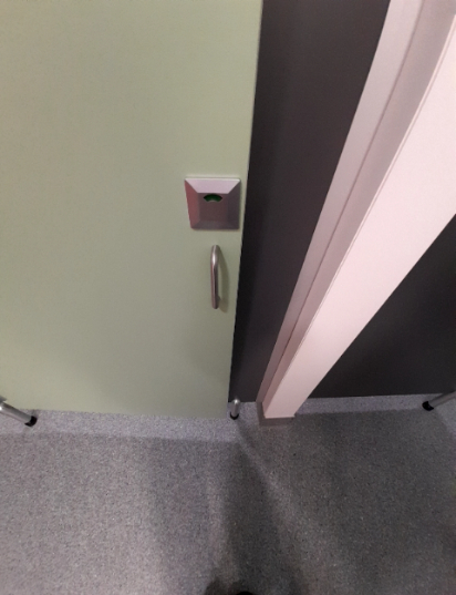 Light green cubicle door with a silver handle and an electronic lock with a green indicator light. The door and an adjacent dark grey partition stand on silver posts above a level, speckled grey fl...