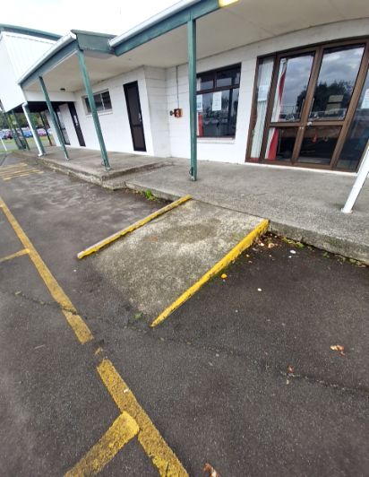 Concrete ramp with yellow edges leads from an asphalt car park to a level concrete verandah. Green pillars frame a glass double-door entrance set in white block walls.