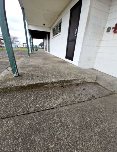 Textured concrete walkway with a single step up to a raised porch area. White block building wall with a black door and dark green metal support posts.