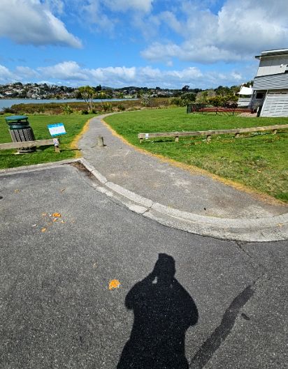Level bitumen path with yellow edges leads from a carpark. A low wooden bollard sits in the centre of the path entrance. A green rubbish bin and wooden rail fence are adjacent.