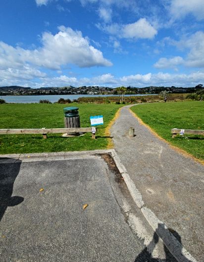 Level gravel path leads from asphalt, through a gap in low wooden fences with a central bollard. Green-lidded rubbish bin and small blue sign on grass to the left. Water in distance.
