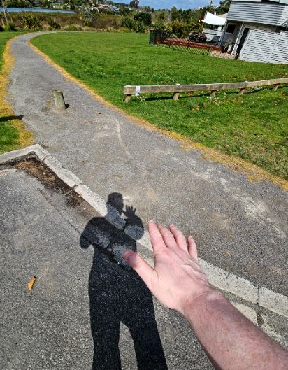 Wide gravel path with an uneven surface curves through green grass. Foreground is level concrete. A low wooden bollard sits on the left. A timber post-and-rail fence is to the right.