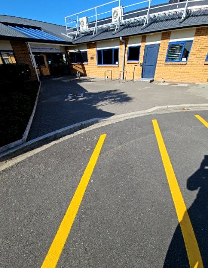 Asphalt car park with yellow lines leads to a level paved path and step-free entrance. Tan brick building features blue door frames and metal racks on the wide, level paved frontage.