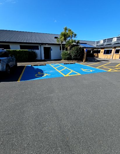 Two level, blue-painted accessible parking spaces with yellow wheelchair symbols and a shared hatched access aisle. Black wheel stops at the head of each bay, near a white building entrance.