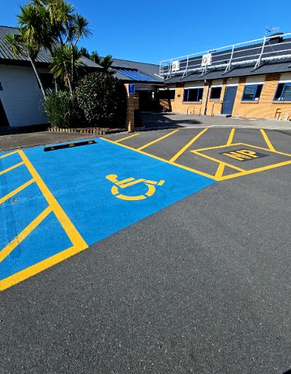 Blue-painted parking bay with yellow wheelchair symbol and hatch markings on level asphalt. A small blue sign is on a brick pillar near the level building entrance.