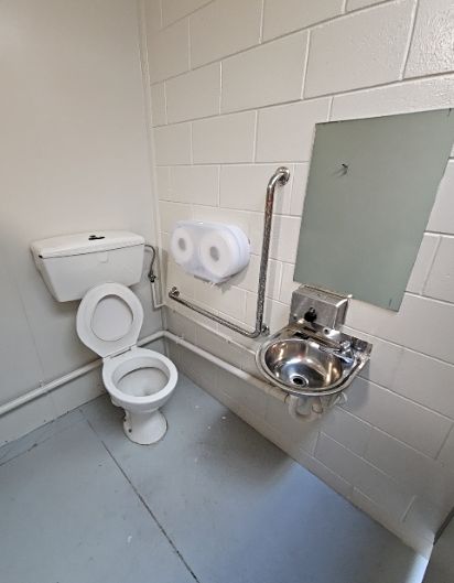 White toilet with L-shaped stainless steel grab rail and small wall-mounted stainless steel sink. Grey smooth floor and off-white concrete block walls with exposed piping.