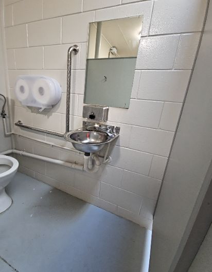 Toilet room with stainless steel L-shaped and horizontal grab rails. Small wall-mounted stainless steel sink, mirror, and white toilet. Level grey floor and white painted block walls.