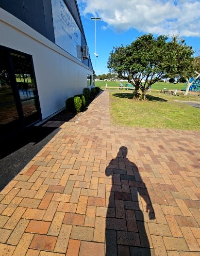 Wide level brick-paved pathway leads to a ground-level glass door entrance. Reddish-brown pavers provide a flat, unobstructed surface alongside a lawn and light-coloured building.