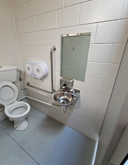 Compact bathroom with white block walls and grey floor. Features a toilet with silver L-shaped grab rail, wall-mounted stainless steel washbasin, mirror, and exposed pipes along the wall base.