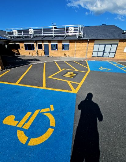Two level accessible parking bays painted blue with yellow symbols. A yellow hatched 'NP' zone separates the bays on asphalt in front of a brick building with a dark blue door.