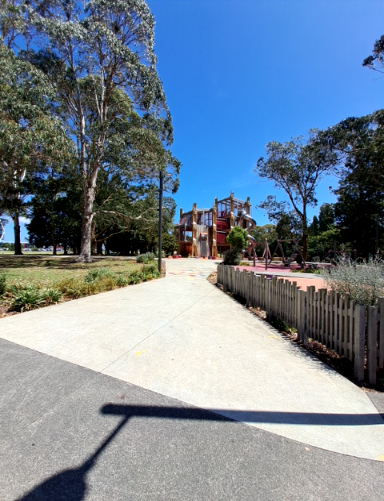 Wide, level concrete path transitioning from asphalt, leading directly to a large multi-level playground structure. A wooden picket fence borders the path on the right.