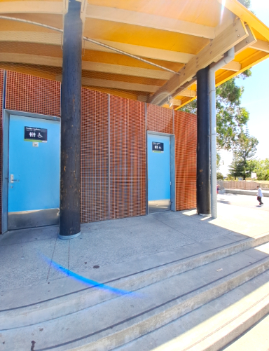 Public toilets with two light blue doors; one general/accessible, one accessible. Under a yellow shelter, a concrete landing has concrete stairs descending in the foreground.