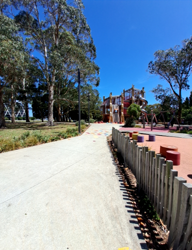 Wide, level concrete pathway with colourful squares leads through a park to a playground with a large structure and softfall. A low wooden picket fence borders the path on the right.