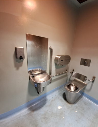 Stainless steel basin and toilet with grab bars. Stainless steel mirror, soap dispenser, and paper towel dispenser. Level, worn light blue-grey floor with blue cove base.