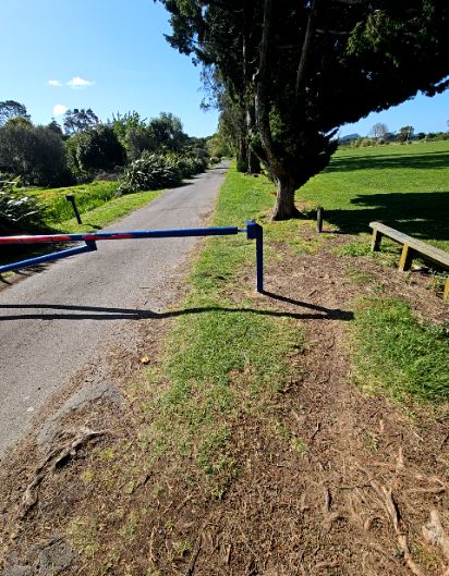 Level asphalt path blocked by a blue metal boom gate. A narrow, uneven bypass path of dirt and grass with exposed tree roots leads around the gate post towards a wooden bench on grass.
