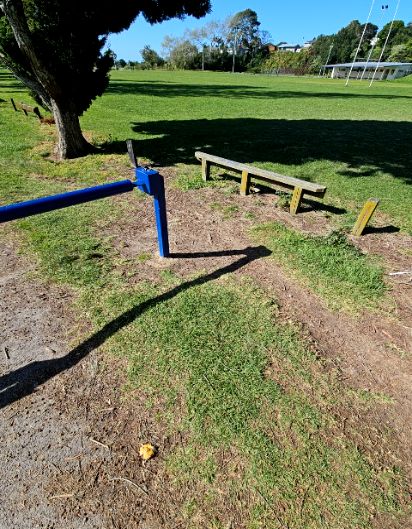 Blue metal swing gate and wooden bench on uneven dirt and patchy grass. Beyond is a level, wide green grass field with sports facilities in the background.