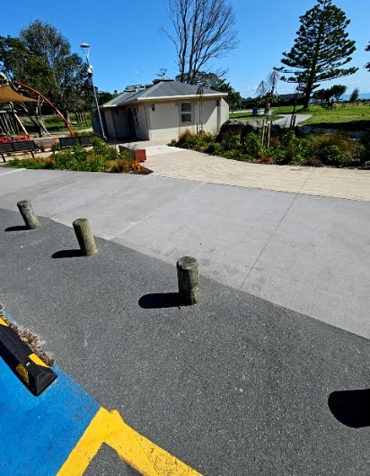 Wide, level asphalt and concrete pathways lead to a beige building. Three wooden bollards border a blue and yellow marked parking space. Surfaces are flat and step-free.