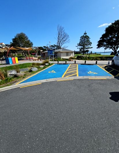 Two blue accessible parking spaces with yellow wheelchair symbols on level asphalt. A concrete kerb borders the spaces, with a playground and light-coloured building in the background.
