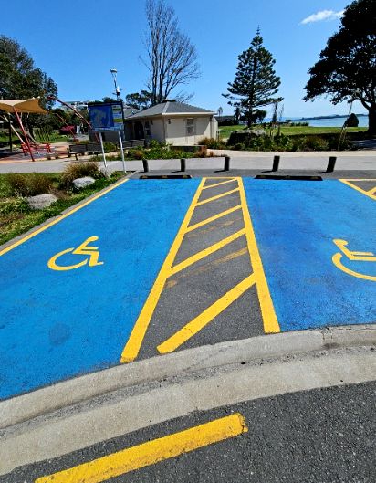 Two blue-painted accessible parking bays with yellow wheelchair symbols and a diagonal-striped buffer. Level asphalt surface with a low concrete kerb and nearby paved park pathway.