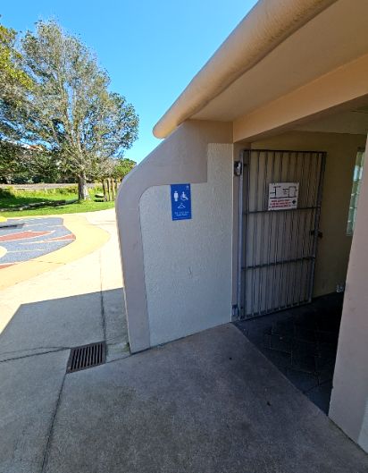 Level concrete pathway with a small drainage grate leads to a wide entrance with an open grey metal gate. A blue sign on the wall features white icons for a person, wheelchair, and clothes hanger.