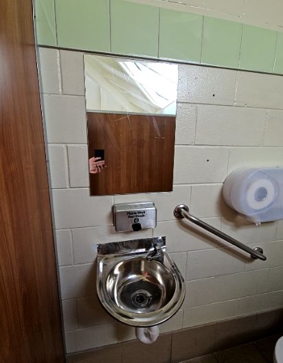 Wall-mounted stainless steel sink with soap dispenser and mirror above. An angled metal grab rail is fixed to the right on light-coloured tiled walls.