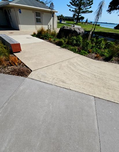 Wide level concrete pathways in grey and tan lead toward a building. A long timber bench with a bright orange end sits beside the path. Flat garden beds and a large rock border the walkway.