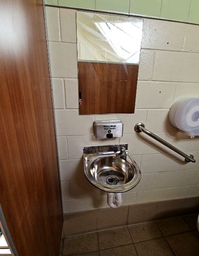 Wall-mounted stainless steel sink with soap dispenser and mirror above. To the right is an angled metal grab rail and toilet roll dispenser on cream tiled walls. Brown tiled floor.