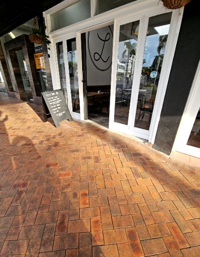 Level entry with white-framed glass doors and open doorway. Wide reddish-brown herringbone brick-paved sidewalk. Black A-frame chalkboard sign to the left of the entrance.