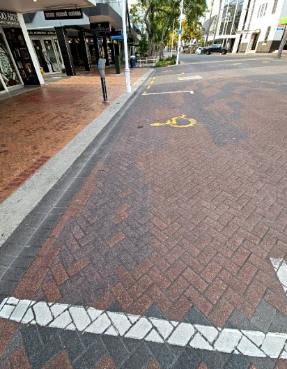 Accessible parking space marked with a yellow wheelchair symbol on herringbone brick paving. A white line borders the space next to a level grey kerb and a wide brick footpath with shopfronts.