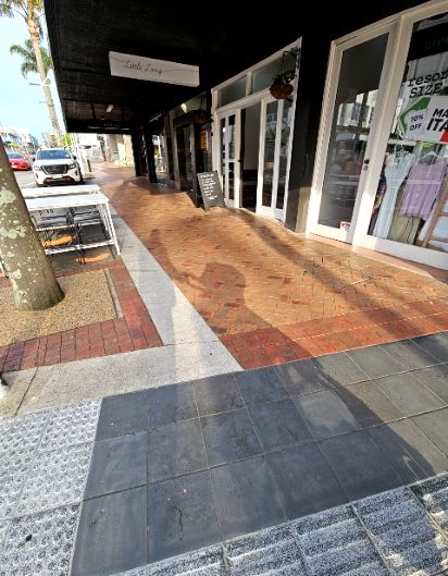 Level footpath with dark grey tiles, tactile paving and reddish-brown bricks. Wide path under a dark awning leads to level shop entrances. Outdoor tables and a sign board are on the left side.