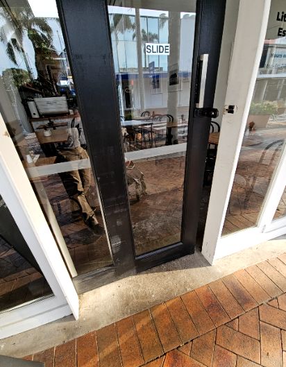 Sliding glass door with black frame and vertical silver handle. White 'SLIDE' sign on glass. Level concrete threshold from reddish-brown brick paving to a tiled floor with indoor seating.