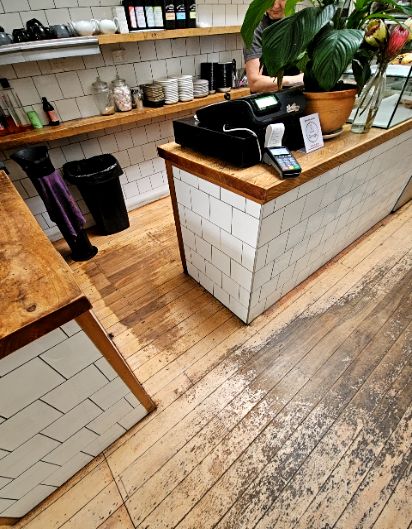 Level light-coloured timber floor with dark worn patches leading to a high timber-topped counter with white tiled sides. Cash register and EFTPOS terminal are on the counter surface.