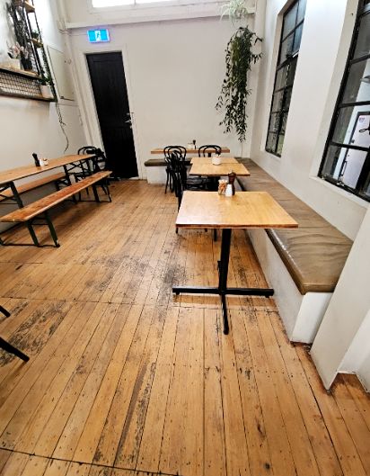 Level light-coloured timber floorboards. Wooden tables on black pedestals with black chairs and a cushioned bench. Wide pathways lead to a black door at the rear with an exit sign above.