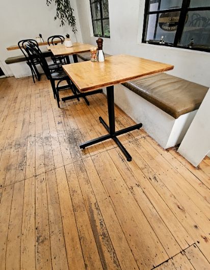 Level light-coloured wooden floorboards. Square wooden tables on black pedestal bases. Seating includes black chairs and a built-in wall bench with a brown padded cushion.