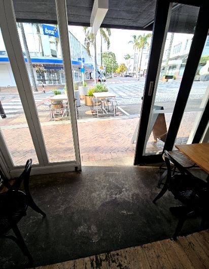 Level threshold entry through wide glass doors from a dark concrete floor to red brick paving. Outdoor wooden tables and metal stools sit on a flat, even surface. Blue BNZ sign across the street.
