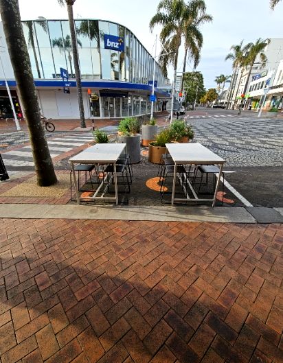 Red-brown herringbone brick walkway leads past a small concrete kerb to two outdoor tables and stools. A level pedestrian crossing leads to a glass-fronted bank building with blue signage.