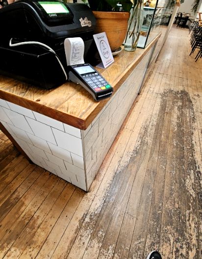 Level timber floorboards with worn patches run alongside a white tiled counter with a wooden top. The floor provides a flat, clear pathway past the service area towards indoor seating.