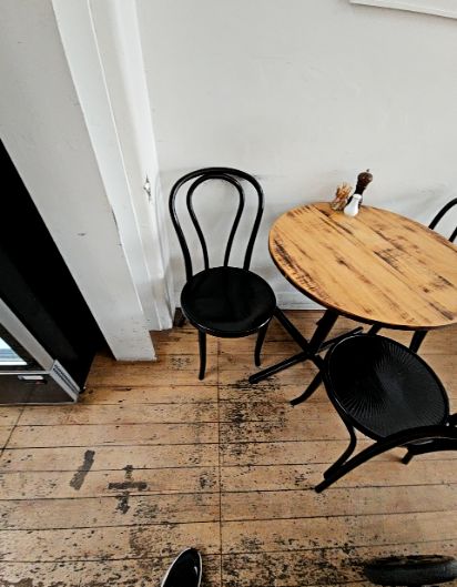 Level, scuffed timber floorboards with a round wooden table on a black metal base. Two black bentwood-style chairs provide seating against a white wall.