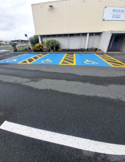 Three blue accessible parking bays with yellow wheelchair symbols and diagonal stripes, on a dark asphalt surface in front of a building.