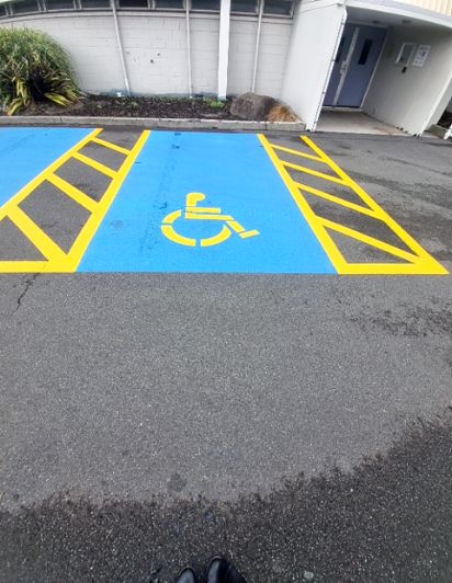 Accessible parking bay on asphalt, painted blue with yellow wheelchair symbol and hatched access aisles on both sides. Building entrance nearby.