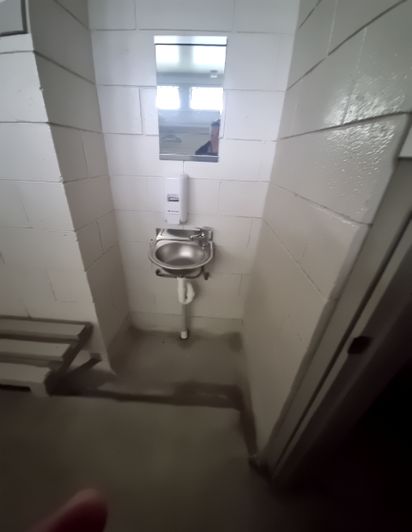 Narrow white-walled utility space with a stainless steel sink, soap dispenser, and mirror. Dark, wet concrete floor. Plank bench on left, door frame on right.