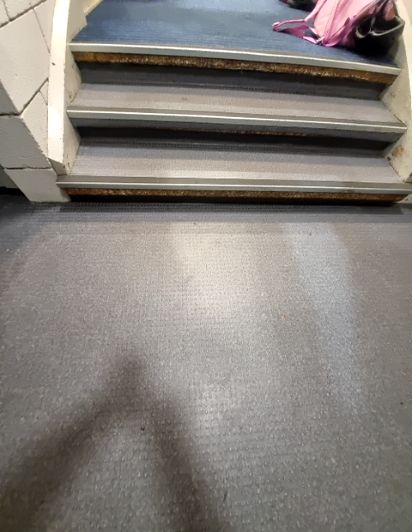 Three steps with grey carpeted treads, dark risers, and silver nosings. Rough brown material is exposed under each nosing. No handrails are visible. Grey textured floor below; a pink bag is on the ...