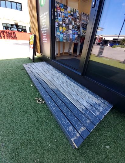 Low, wide dark blue composite ramp with a gentle gradient from artificial turf to a glass-doored entrance. The ramp surface shows wear. An A-frame sign is to the left of the door.