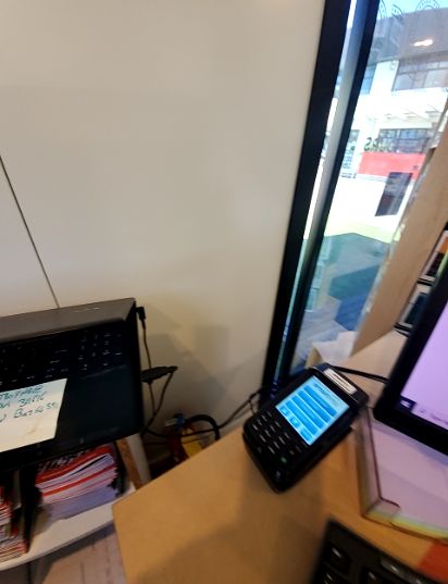Standard-height light brown desk with a black payment terminal with an illuminated screen, computer monitor, and keyboard. A large window provides natural light. A shelf with magazines is beneath t...