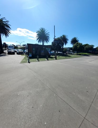 Wide grey concrete forecourt leading to a paved area with parked cars on the left. A small dark building stands on artificial grass in the centre with bollards.