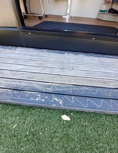 Raised grey composite decking with horizontal ridges and white scuff marks leads to a black doorway threshold. Green artificial grass below.